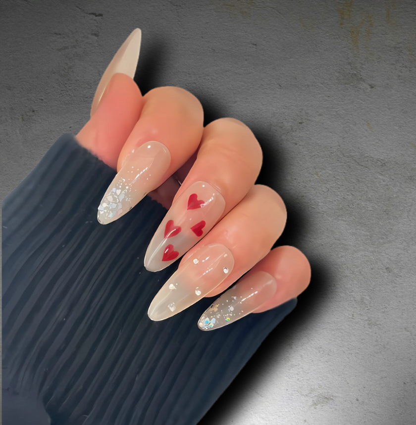 long almond-shaped press-on nails featuring a nude base with red hearts, subtle glitter accents, and a glossy finish.
