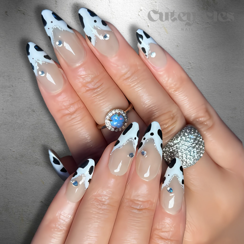 Cow-print almond nails with black-and-white design and rhinestone accents on a nude base.