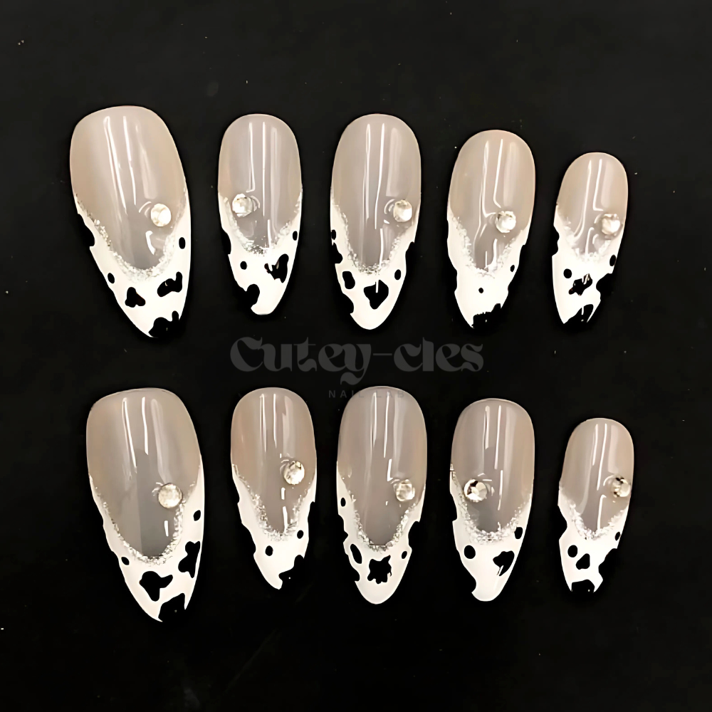 Cow-print almond nails with black-and-white design and rhinestone accents on a nude base.