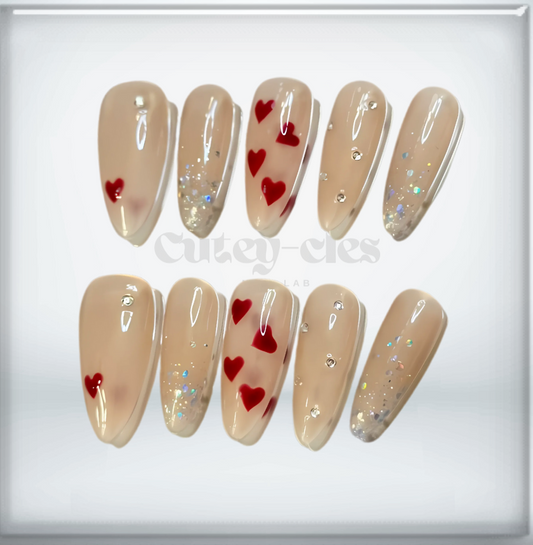 long almond-shaped press-on nails featuring a nude base with red hearts, subtle glitter accents, and a glossy finish.