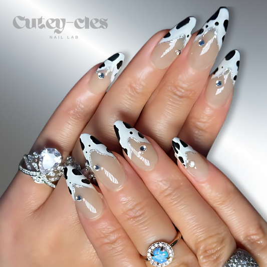 Cow-print almond nails with black-and-white design and rhinestone accents on a nude base.