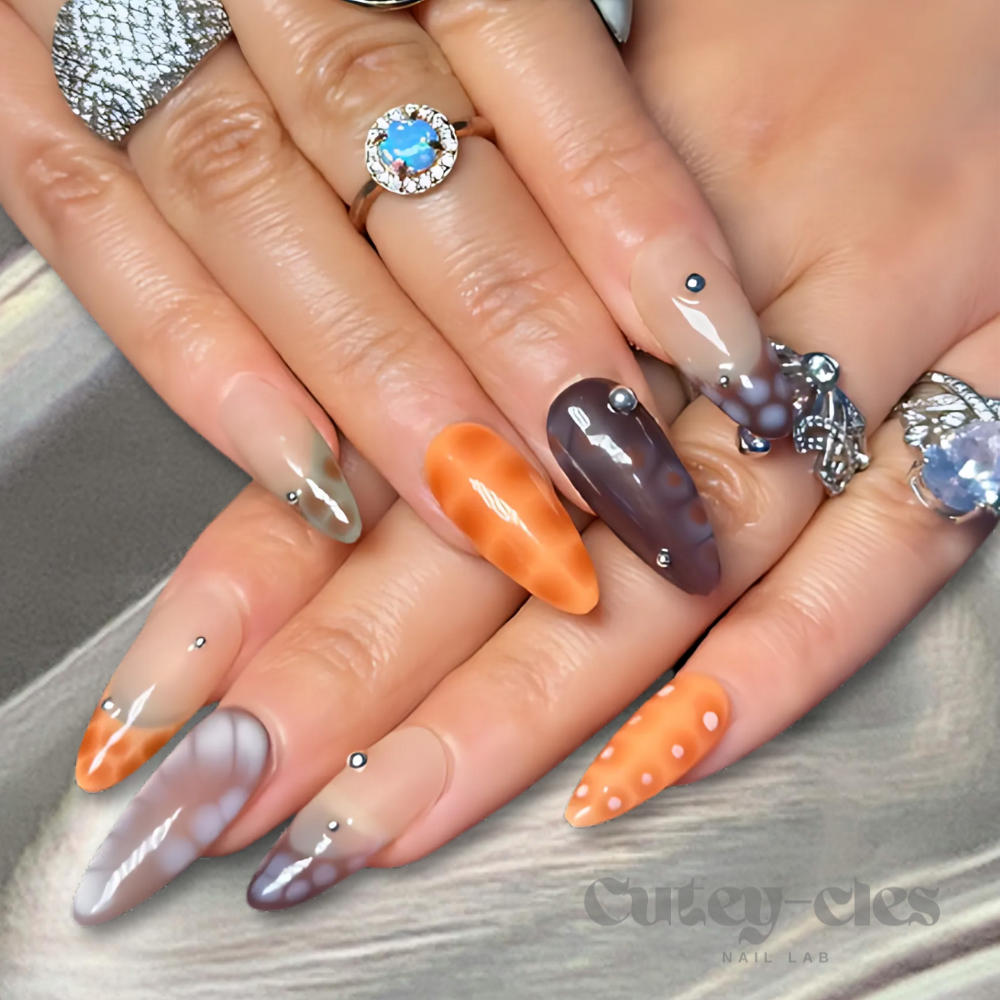 Medium almond press-on nails with a fall-inspired abstract design featuring warm earthy tones, metallic accents, and 3D embellishments, displayed on a model's hands and on a decorative background.