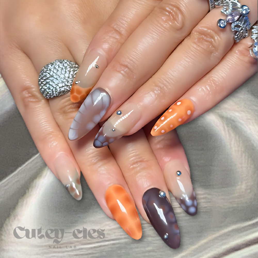 Medium almond press-on nails with a fall-inspired abstract design featuring warm earthy tones, metallic accents, and 3D embellishments, displayed on a model's hands and on a decorative background.