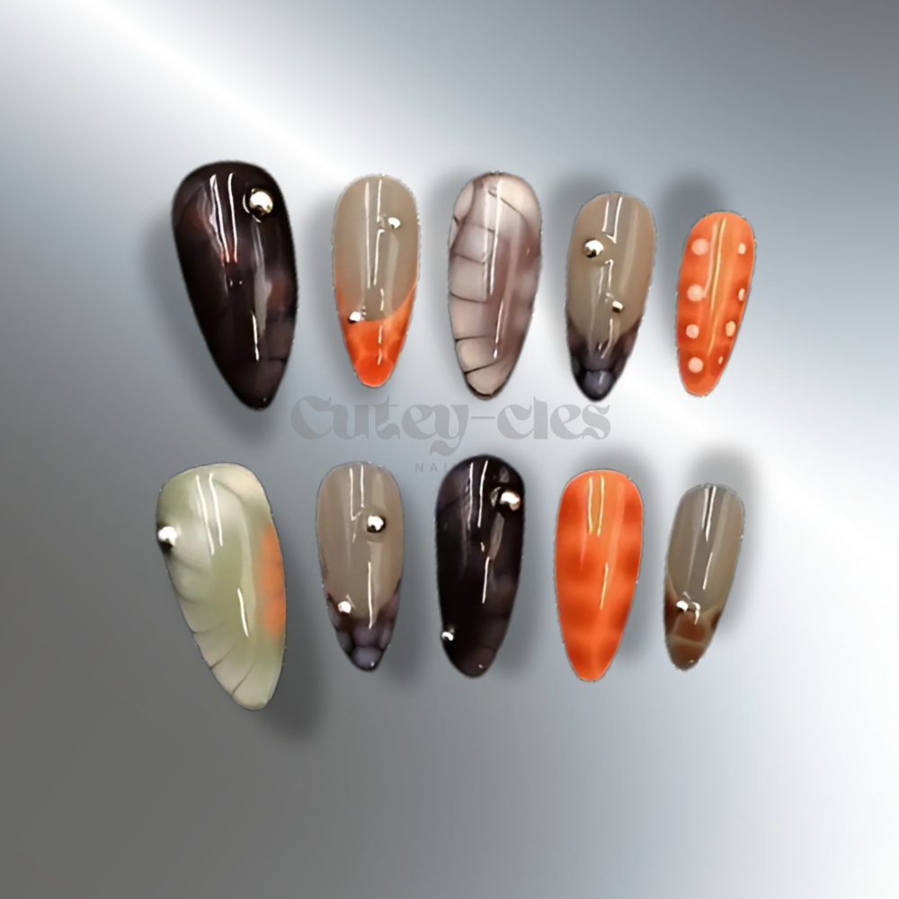 Medium almond press-on nails with a fall-inspired abstract design featuring warm earthy tones, metallic accents, and 3D embellishments, displayed on a model's hands and on a decorative background.