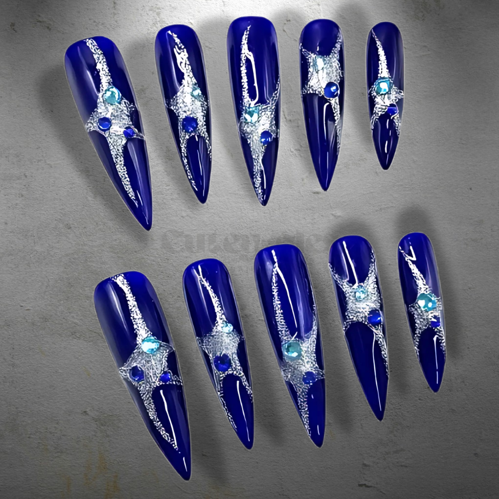 A set of cobalt blue stiletto nails with silver starburst designs and sparkling gemstones.