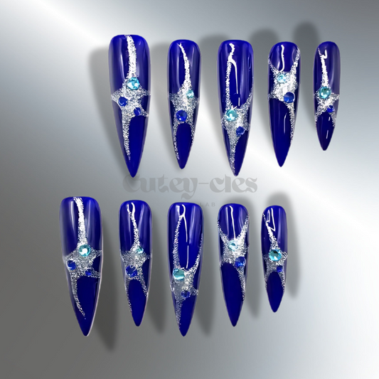 A set of cobalt blue stiletto nails with silver starburst designs and sparkling gemstones