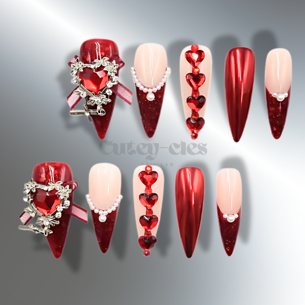 Valentine’s Day-themed press-on nails in deep red with heart-shaped rhinestones, pearl details, and glossy metallic finishes.