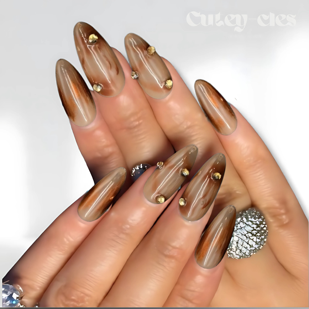 Elegant almond-shaped press-on nails with a brown marble design, glossy finish, and gold stud accents.