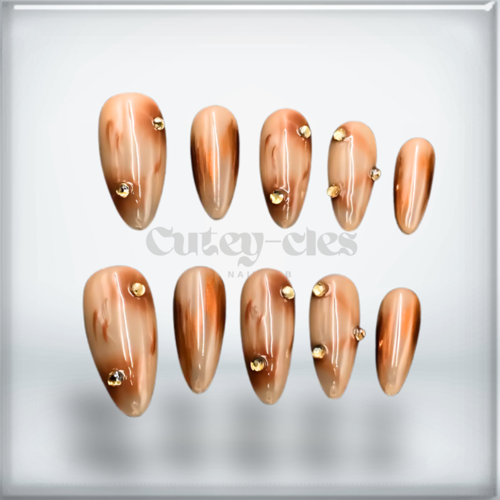 Elegant almond-shaped press-on nails with a brown marble design, glossy finish, and gold stud accents.