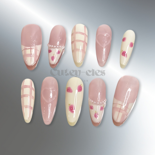 Almond press-on nails with hand-painted strawberry designs, pastel pink grids, glitter accents, and pearl embellishments on a textured background.