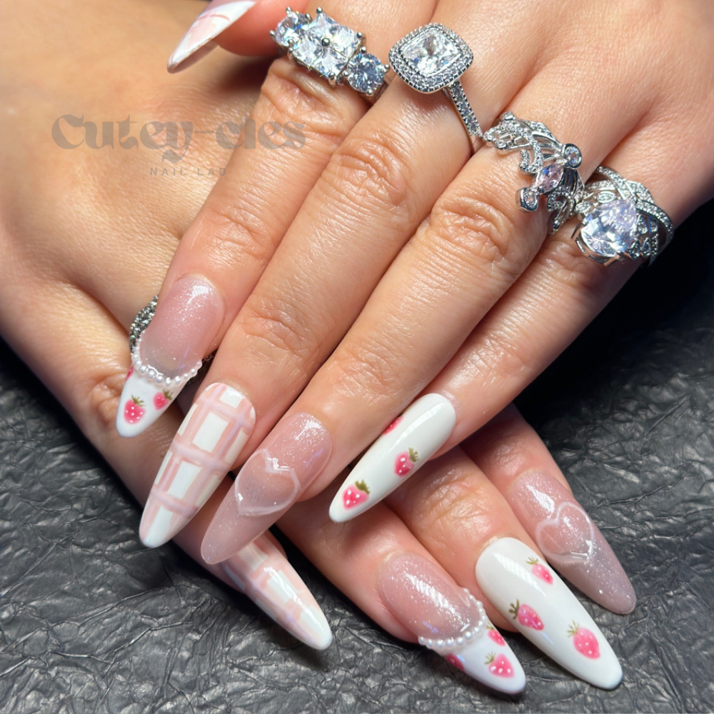 Cool almond press on nails with hand-painted strawberry designs, pastel pink grids, glitter accents, and pearl embellishments on a textured background.