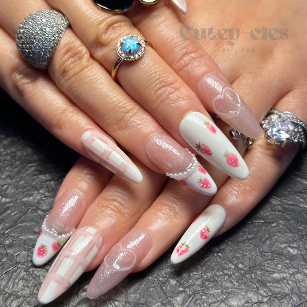 Long almond press-on nails with hand-painted strawberry designs, pastel pink grids, glitter accents, and pearl embellishments on a textured background.